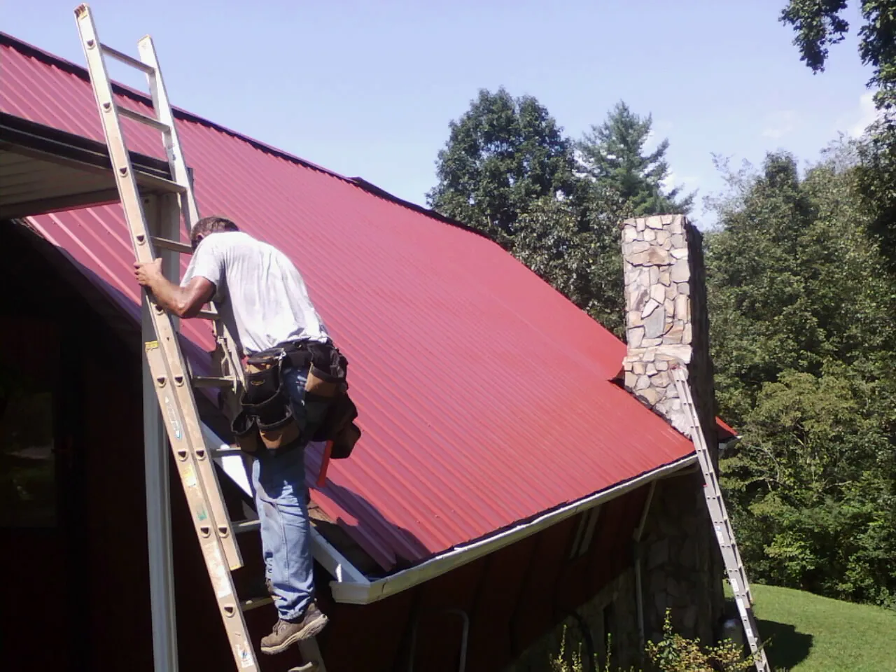 Roofer on a ladder beside a red metal roof and stone chimney — Soffit & Fascia Repair by Roofing Brothers in New Buffalo